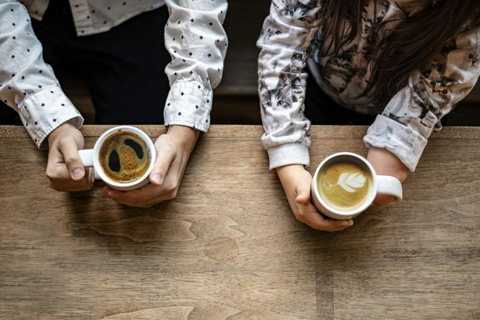 Tripsitter – zwei Personen halten Tassen mit Kaffee in den Händen, ruhiges Gespräch am Holztisch
